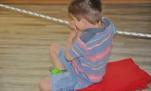 Chłopiec w paski ćwiczy na obniżone napięcie mięśniowe, siedząc na czerwonym kocu.