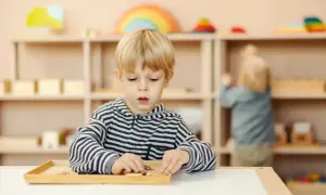 Chłopiec skupiony na zadaniu z materiałami Montessori. W tle inne dziecko i regały z zabawkami.