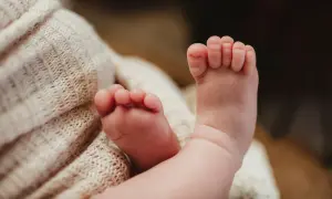 Delikatne, różowe stópki niemowlaka, z widocznymi podwiniętymi palcami u nóg, spoczywają na miękkim, beżowym kocyku.
