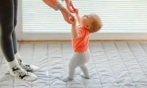 Mama pomaga dziecku, kiedy zaczyna chodzić. Maluch w pomarańczowej bluzeczce i szarych legginsach robi pierwsze kroki.