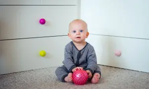 7 miesięczne dziecko z niebieskimi oczami siedzi na dywanie i bawi się różową, kolczastą piłeczką.