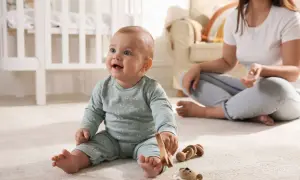 Uśmiechnięte 10 miesięczne dziecko bawi się drewnianymi zabawkami na dywanie, podczas gdy mama siedzi w tle.