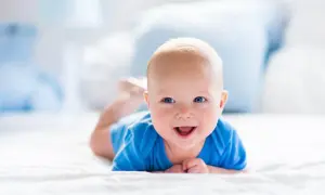 Uśmiechnięty 2 miesięczne dziecko w niebieskim body leży na brzuchu na białej pościeli.