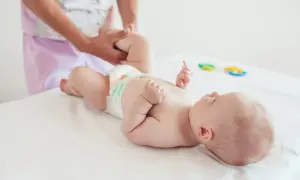 Dziecko leży na białym podłożu, a dorosły delikatnie masuje jego nóżkę. To ważny moment, wspierający ostatni skok rozwojowy malucha.