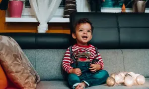 Uśmiechnięty maluch w świątecznym sweterku i ogrodniczkach siedzi na kanapie. Od kiedy można sadzać dziecko z podparciem? Ten maluch już potrafi!