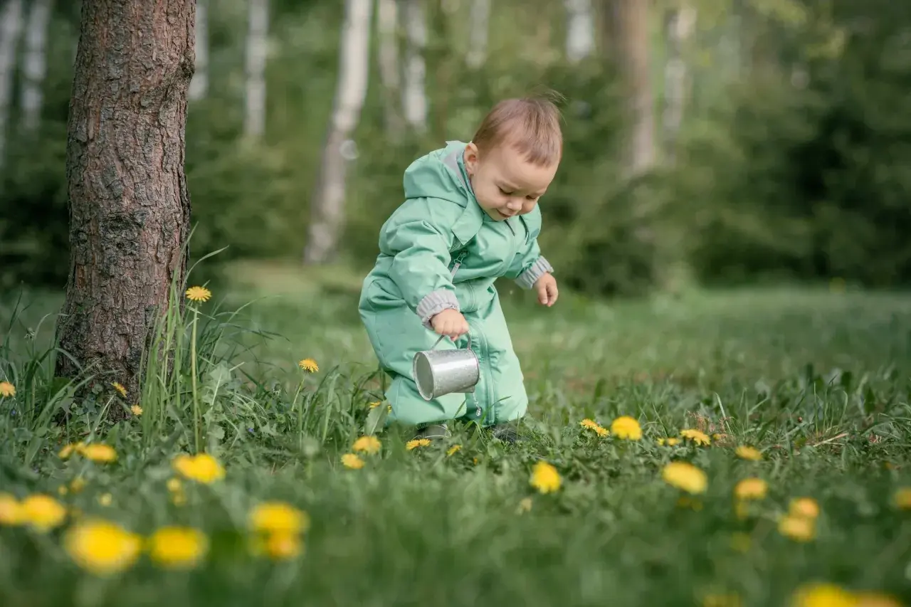 Dziecko w zielonym kombinezonie, z konewką w ręku, odkrywa świat wśród traw i mleczy. To jego **3 skok rozwojowy**, pełen ciekawości i nowych doświadczeń.