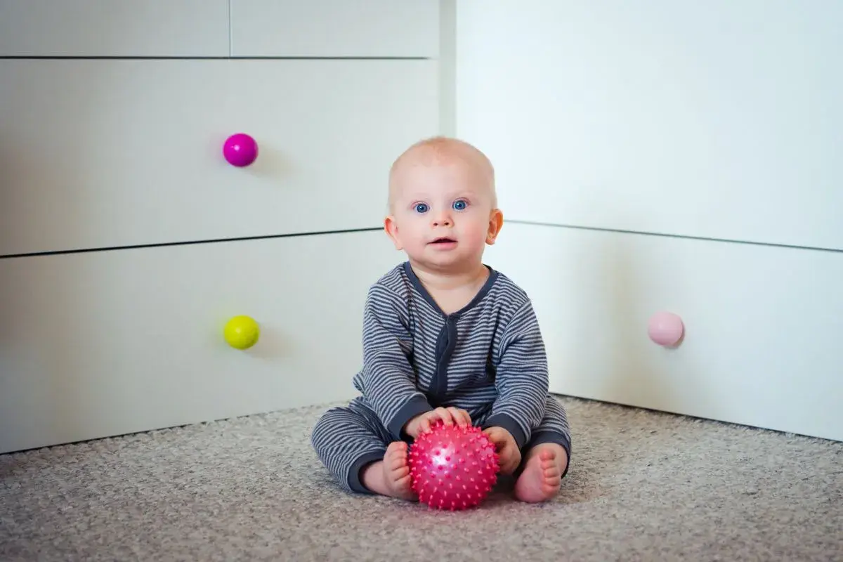 7 miesięczne dziecko z niebieskimi oczami siedzi na dywanie i bawi się różową, kolczastą piłeczką.