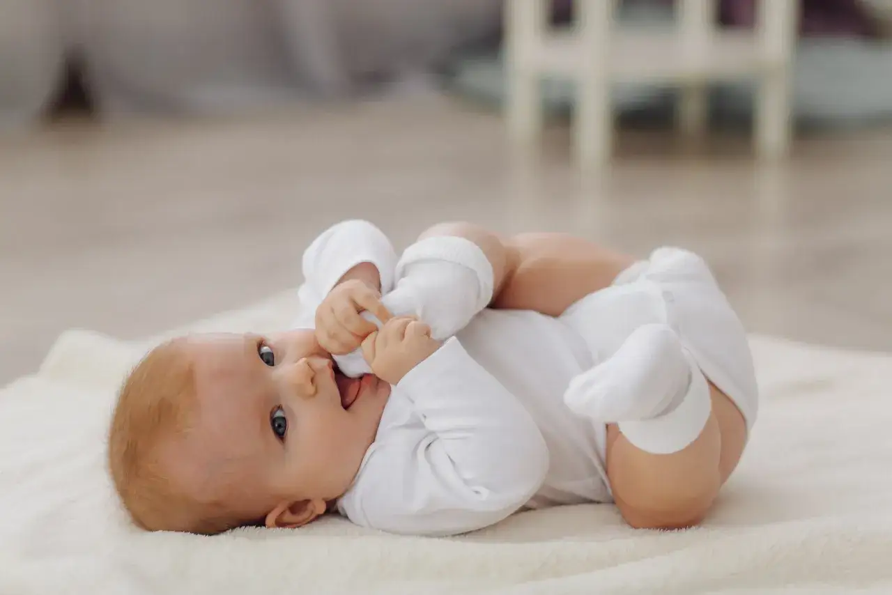 Słodkie, 6 miesięczne dziecko w białym body leży na miękkim kocyku i bawi się swoimi stópkami.