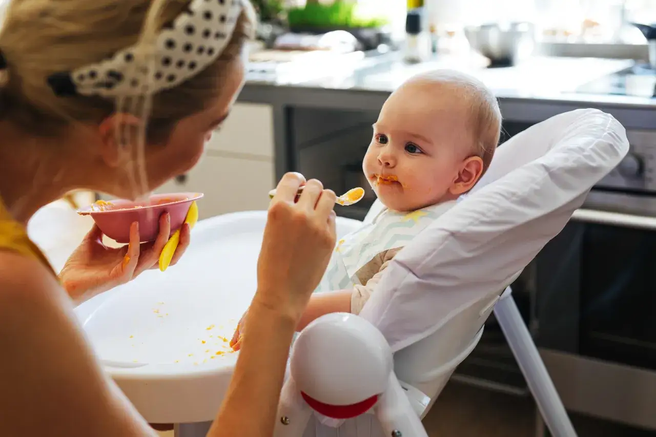 Mama karmi niemowlę, które ma pomarańczowe jedzenie na buzi. To ważny moment, by dowiedzieć się, ile powinno jeść 7 miesięczne niemowlę.