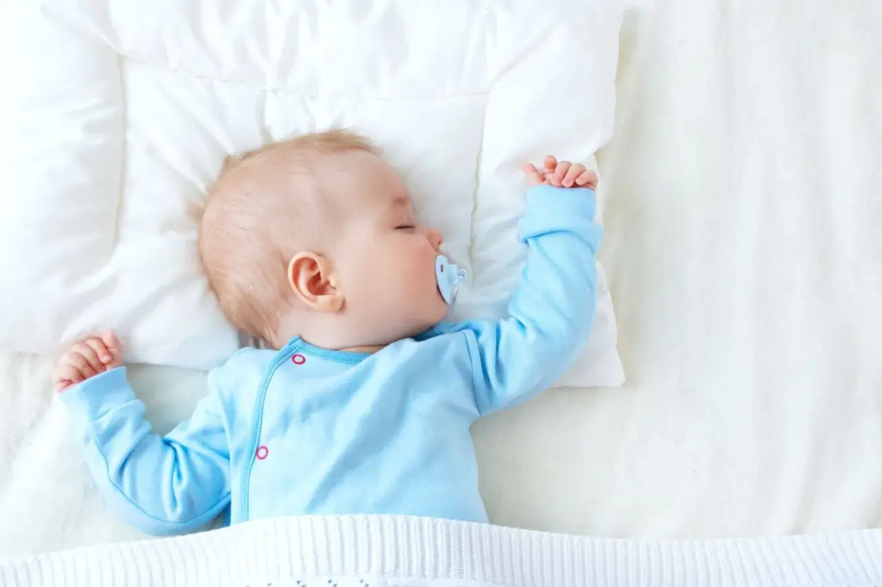 Śpiący niemowlak w niebieskim pajacyku, z niebieskim smoczkiem. Jego rączki ułożone są w lekkiej asymetrii, co może być oznaką asymetrii u niemowlaka.