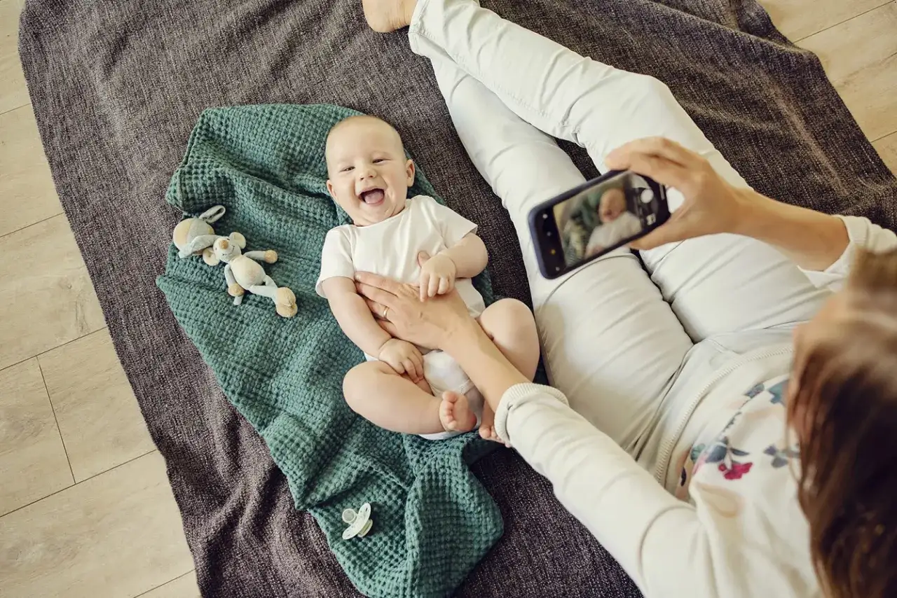 Uśmiechnięte dziecko leży na kocyku, a mama robi mu zdjęcie telefonem. To radosny moment, kiedy dziecko zaczyna wstawać i odkrywać świat.